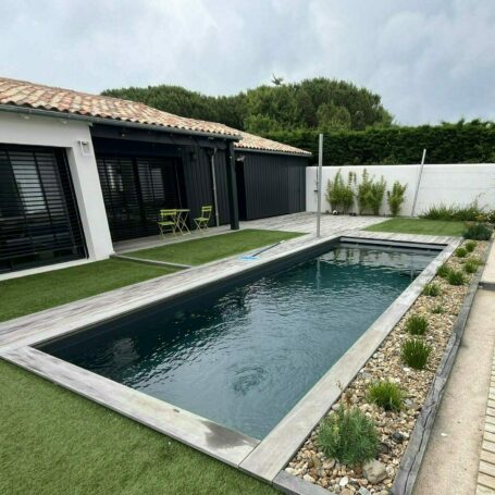 Piscine installée dans le jardin d'un particulier, entourée d'une pelouse verte coupée à ras et d'un aménagement paysager sur le côté droit de la piscine suivi d'une terrasse en bois.