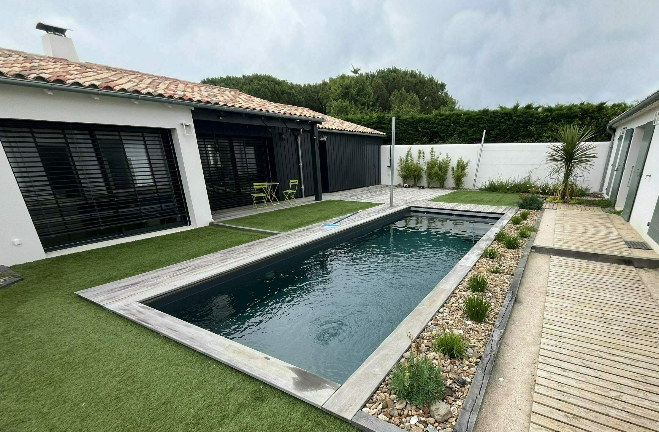 Piscine installée dans le jardin d'un particulier, entourée d'une pelouse verte coupée à ras et d'un aménagement paysager sur le côté droit de la piscine suivi d'une terrasse en bois.